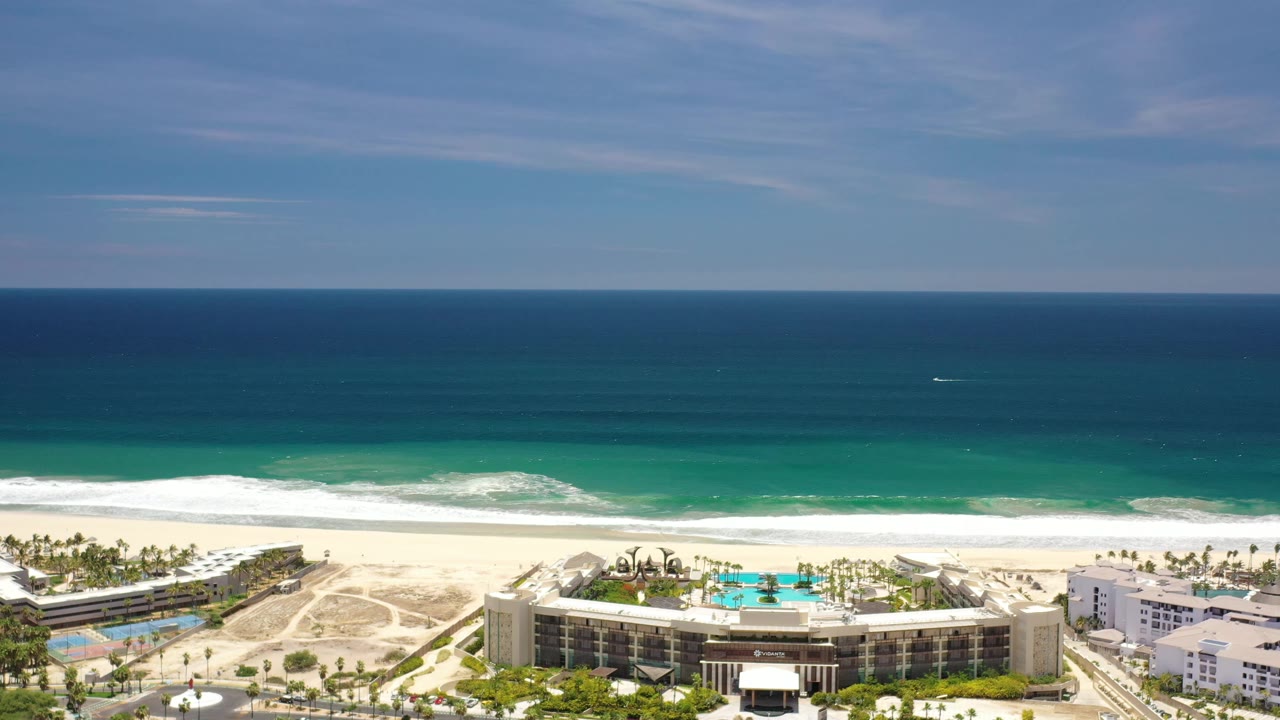 A Los Cabos aerial frame shows layered blue water, a white surf line, pale sand, resort pools, hotel buildings, palms, and no visible people.