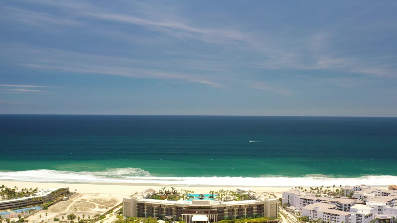 Opening aerial frame over Los Cabos, Mexico, with deep blue ocean on the horizon, turquoise surf near the sand, beachside buildings along the bottom edge, and no visible people.