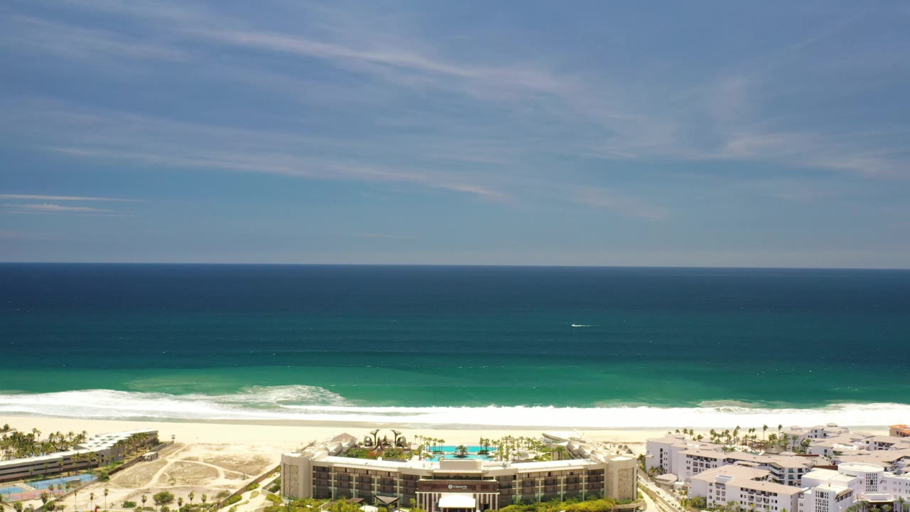 Aerial frame in Los Cabos, Mexico, showing a long ocean horizon, layered blue-green water, white waves reaching the beach, resort buildings at the lower edge, and no visible people.