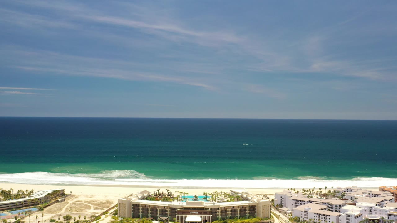 High coastal view with broad sky, dark ocean, surf, beach, and hotel-like buildings.