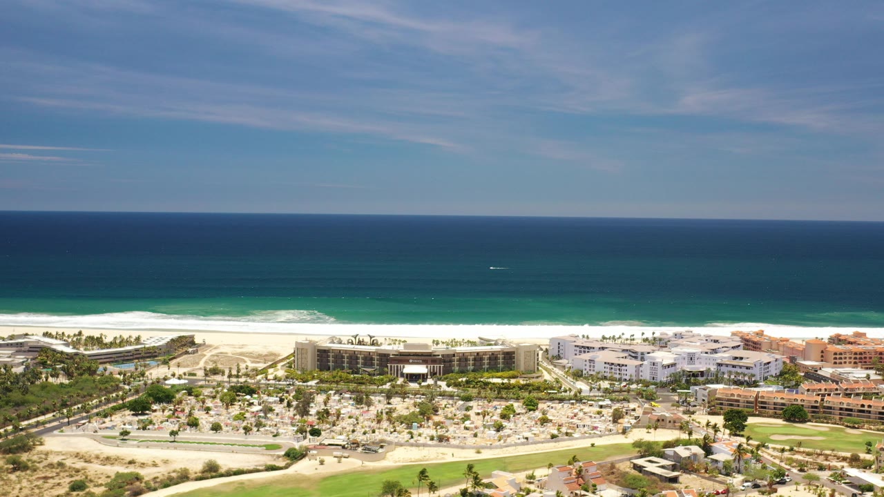 Aerial coastline with blue ocean, beach, palms, roads, and resort buildings.