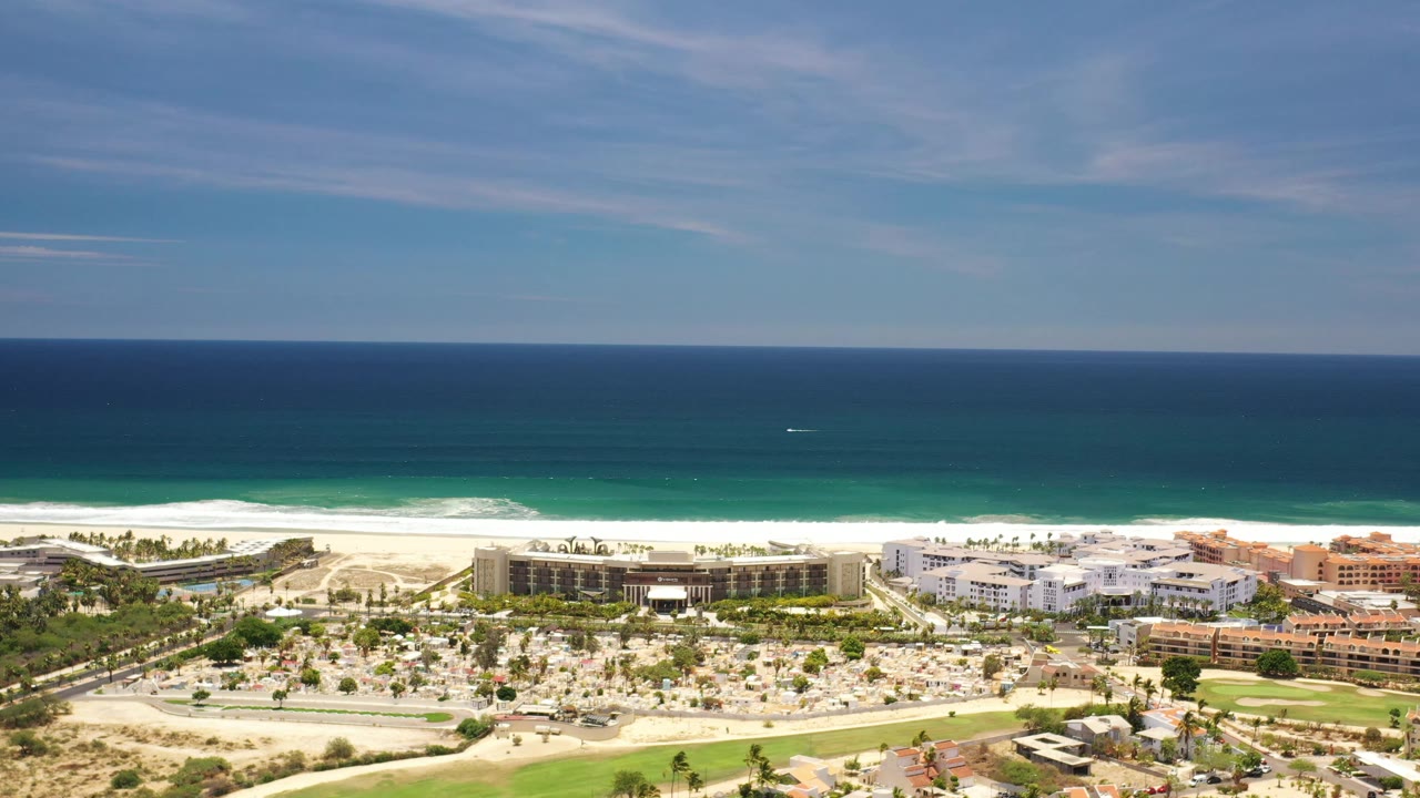 Wide drone-like frame over Los Cabos with the ocean band high in frame, surf on the beach, a line of palms, and low resort buildings below, with no visible people.