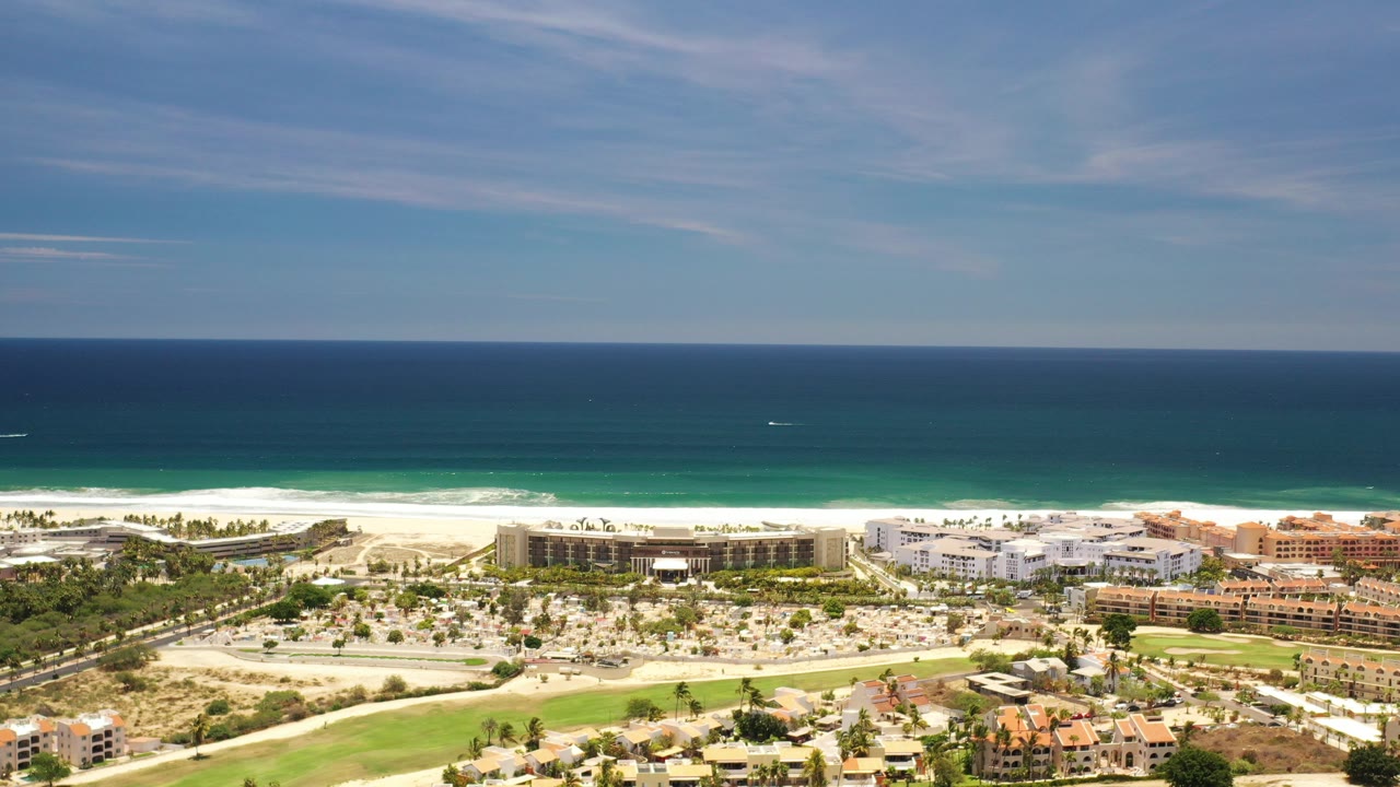 Wide aerial view over Los Cabos, Mexico with blue ocean on the horizon, pale beach, low-rise coastal buildings, roads, green landscaping, and no visible people.