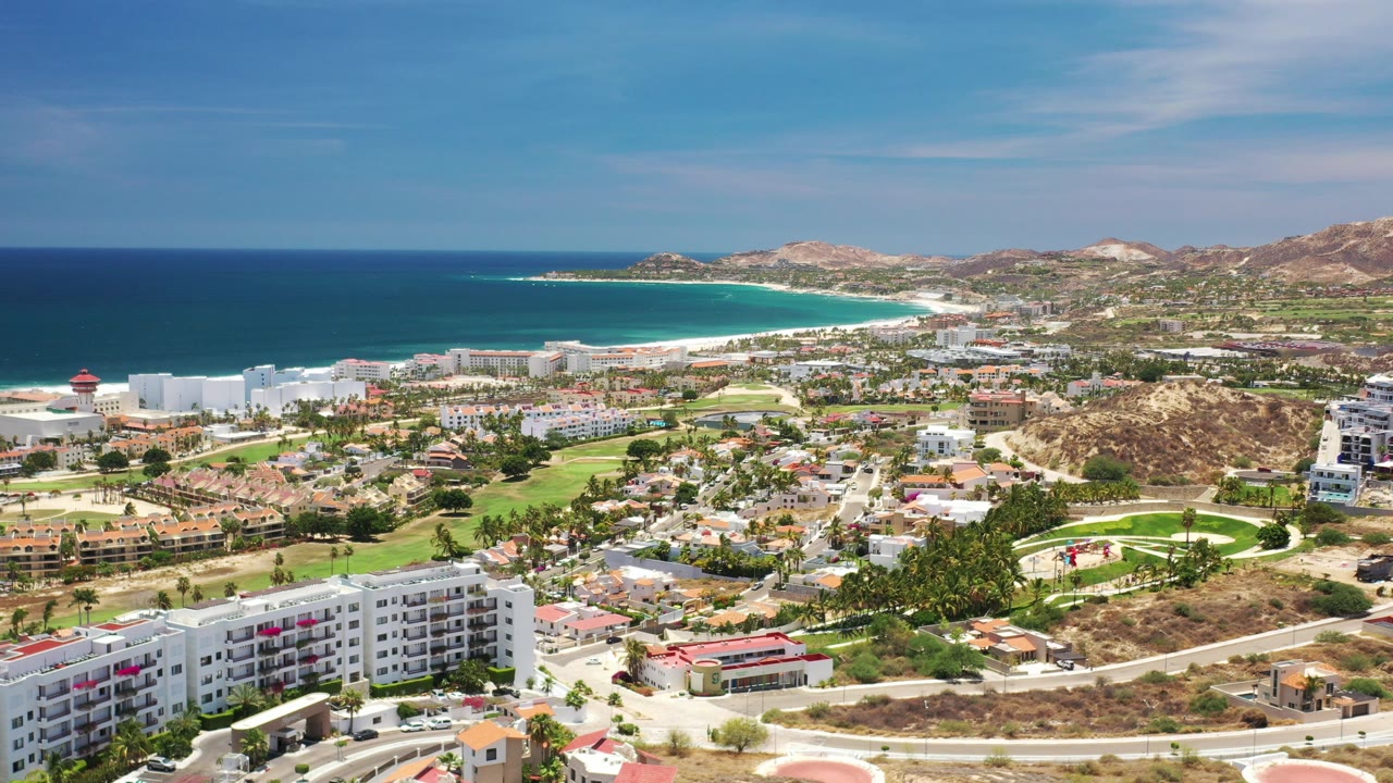 Wide aerial view of a Los Cabos coastal city, beach, ocean, hills, roads, and buildings.