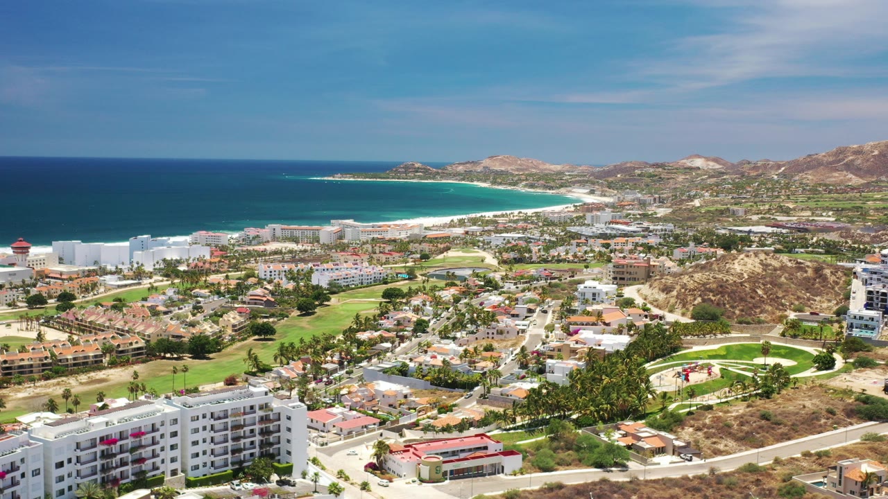 High aerial view over Los Cabos, Mexico, with apartment buildings in the foreground, green fairways and roads through the city, a turquoise bay, dry hills, and no visible people.