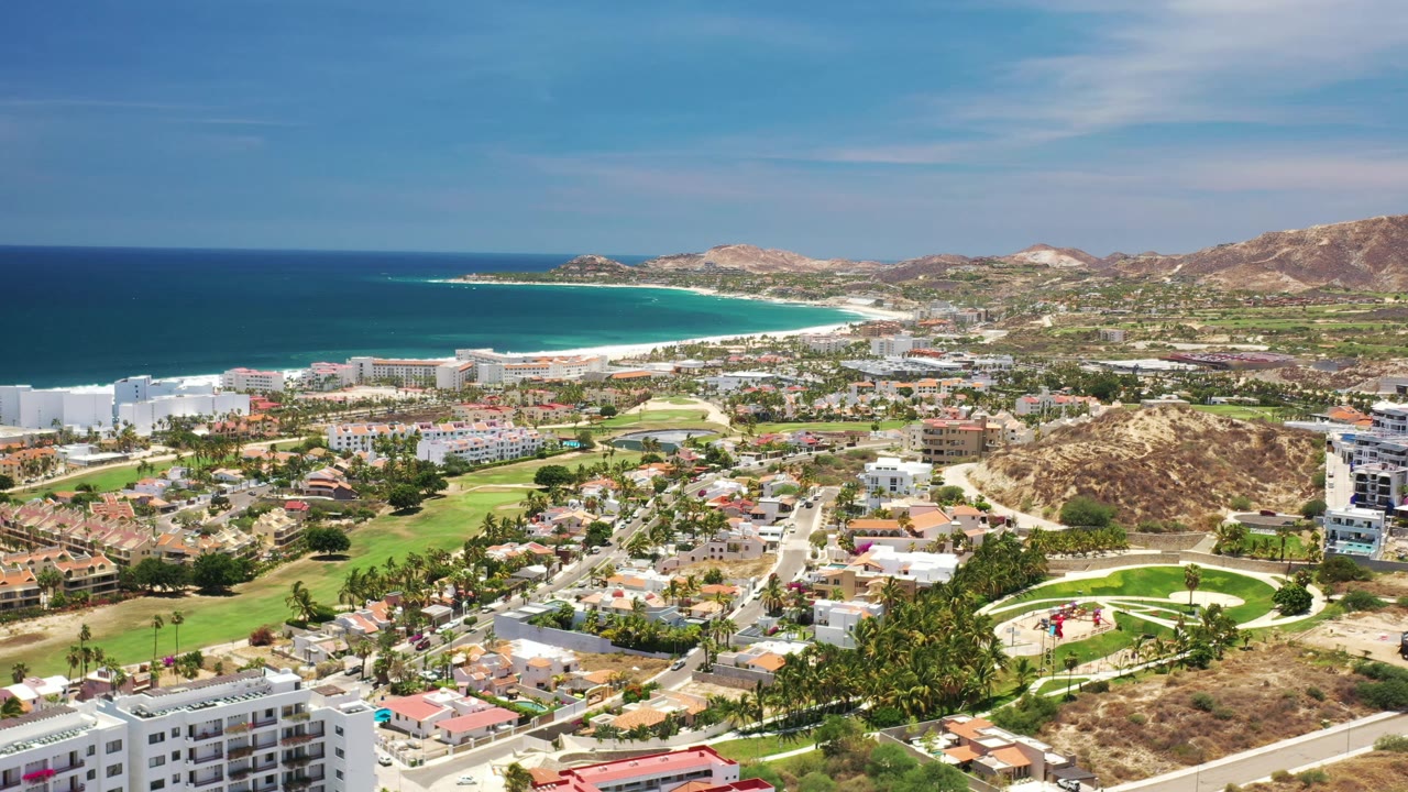 Aerial Los Cabos frame with bright green fairways below, white coastal buildings near the beach, deep blue ocean at left, distant hills, and no visible people.