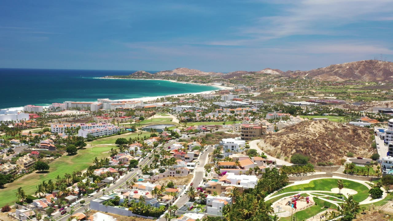 High aerial view over Los Cabos, Mexico with residential streets and green areas in the foreground, a turquoise shoreline to the left, dry hills to the right, and tiny distant people in a park area.