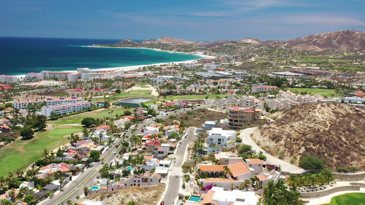 A drone-height view over Los Cabos, Mexico frames foreground villas and roads below golf fairways, hotel blocks, a bright beach, dark blue ocean, and tan hills, with no visible people.