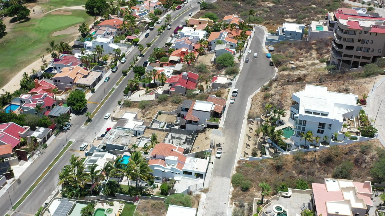 Aerial view in Los Cabos, Mexico, with a white modern house near center, red-roofed homes, a curving side street, dry land, and no visible people.
