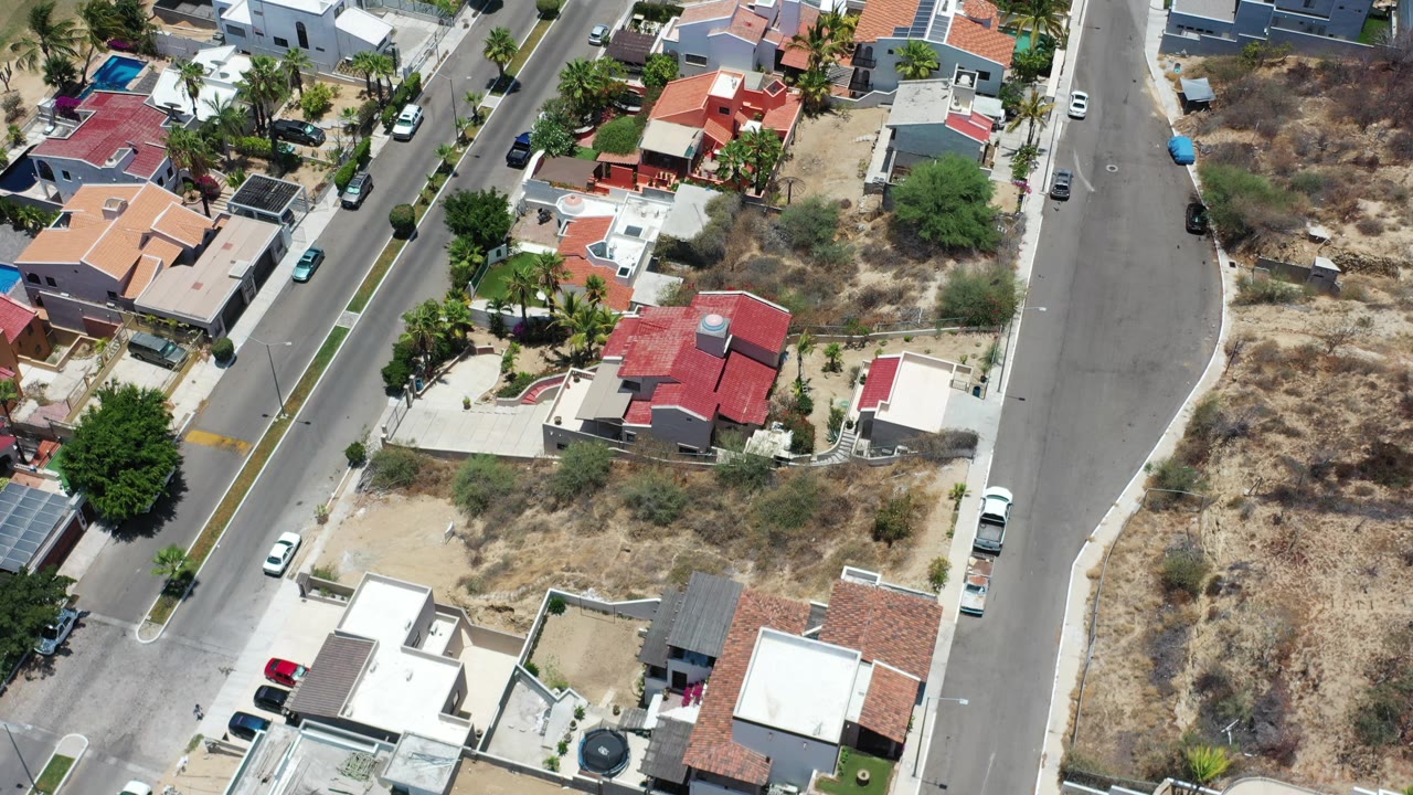 High-angle Los Cabos frame with clustered houses beside a curving road, dry vacant lots, scattered parked cars, palms, and no visible people.