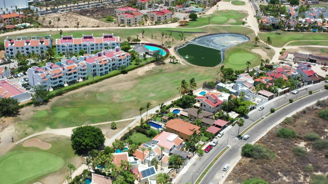 Los Cabos aerial frame with a curving road below, red-roof homes beside green fairways, small blue and dark ponds, and distant hotel buildings near the shore with no visible people.
