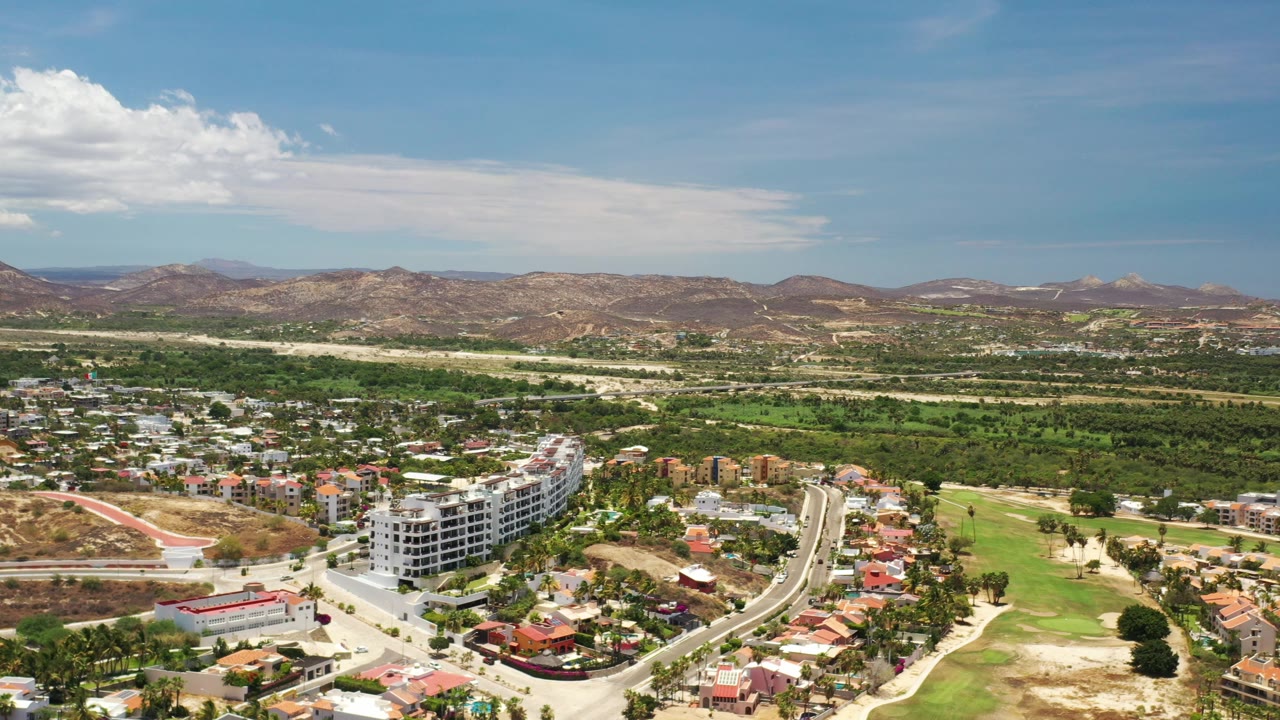 Aerial view in Los Cabos with a curved road, clustered homes, a white multi-story building, green course areas, mountains, and no visible people.