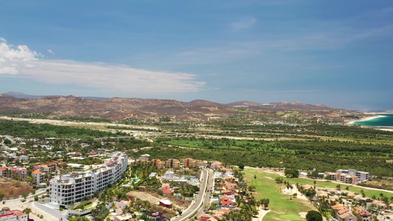 A high Los Cabos aerial frames curving roads, low-rise buildings, green fairway-like land, brown hills, and the turquoise ocean at the right edge, with no visible people.