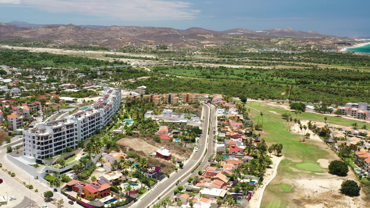 Aerial view of Los Cabos homes beside a road, golf course, hills, and coast.