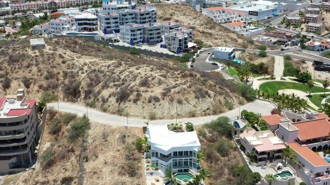 Aerial view in Los Cabos, Mexico, with a scrub-covered slope, a white-roofed house near the lower edge, apartment-style buildings above it, and no visible people.