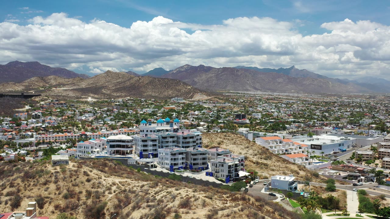 Los Cabos aerial frame with a rounded dry hill at center, residential blocks across the valley, larger white buildings in the foreground, layered mountains behind, and no visible people.