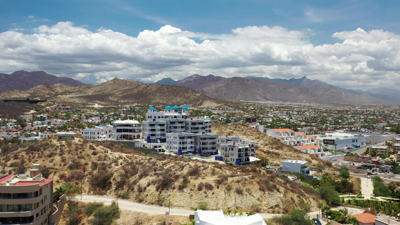 Opening view over Los Cabos, Mexico, with dry hillside terrain, modern white buildings, distant city blocks, mountain ridges, bright clouds, and no visible people.