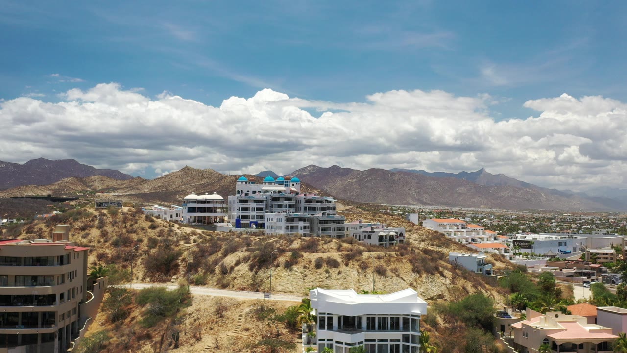 Wide hillside view with modern buildings, mountains, blue sky, and white clouds.