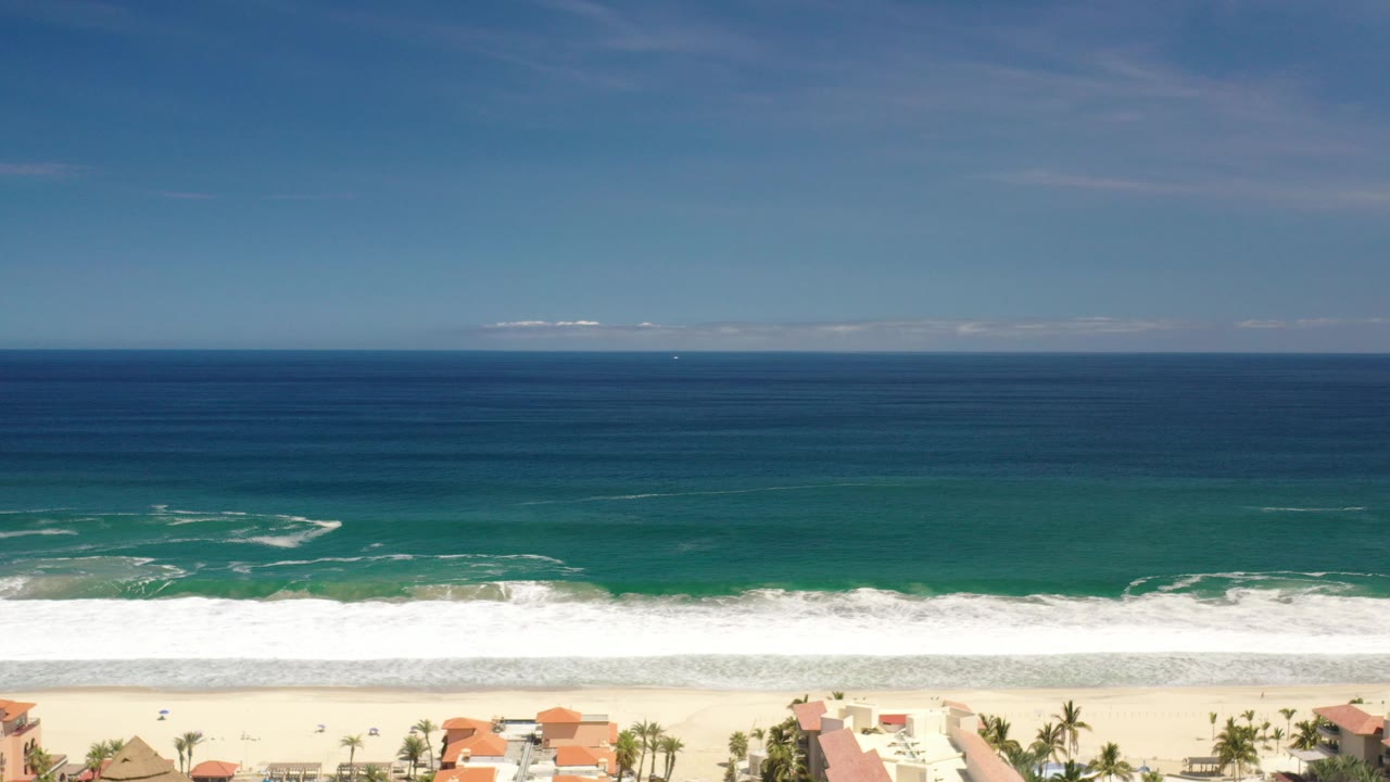 Wide aerial view in Los Cabos, Mexico, with blue ocean bands, white surf, pale sand, and no visible people at the start of the clip.