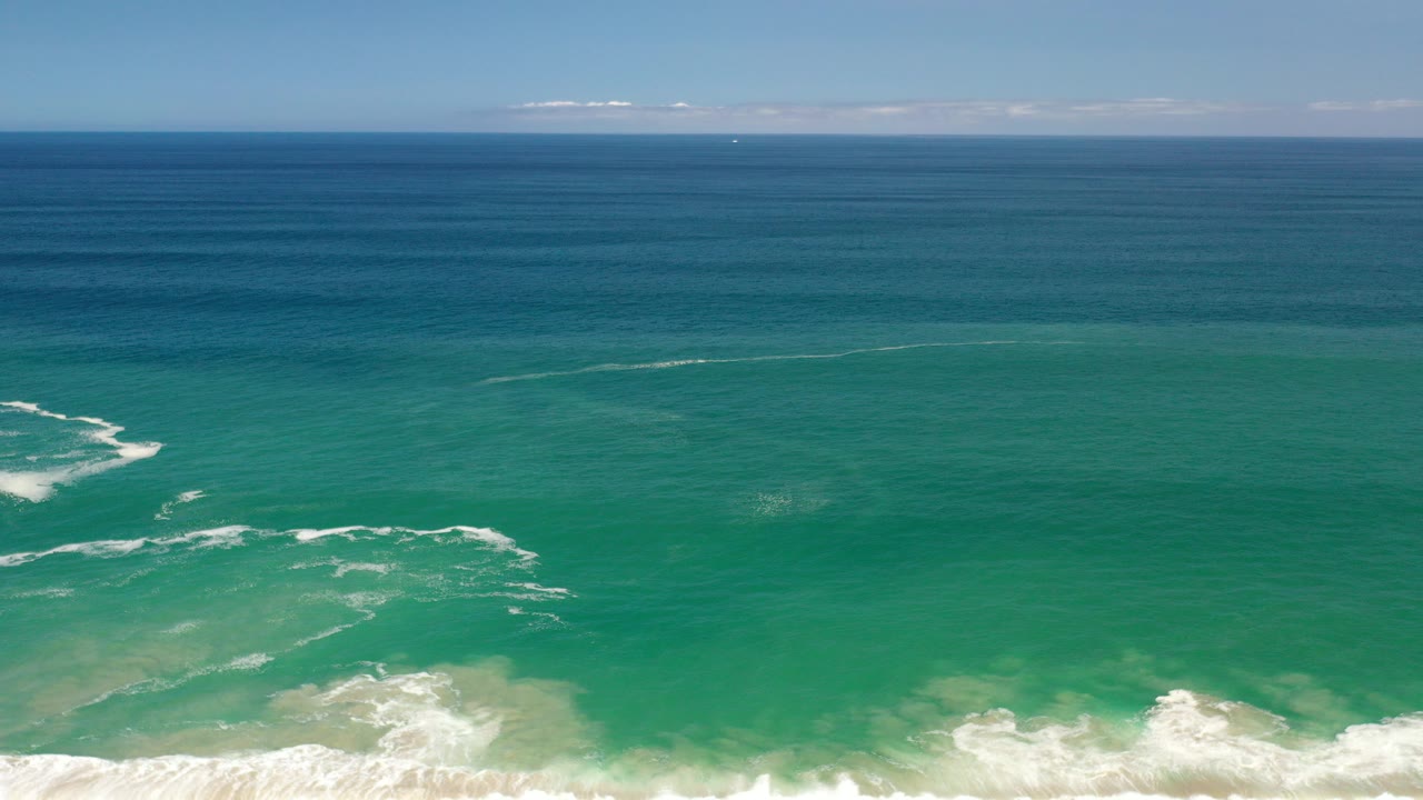 Aerial-looking view over Los Cabos ocean water with turquoise shallows, white surf at the lower edge, a blue horizon, and no visible people.