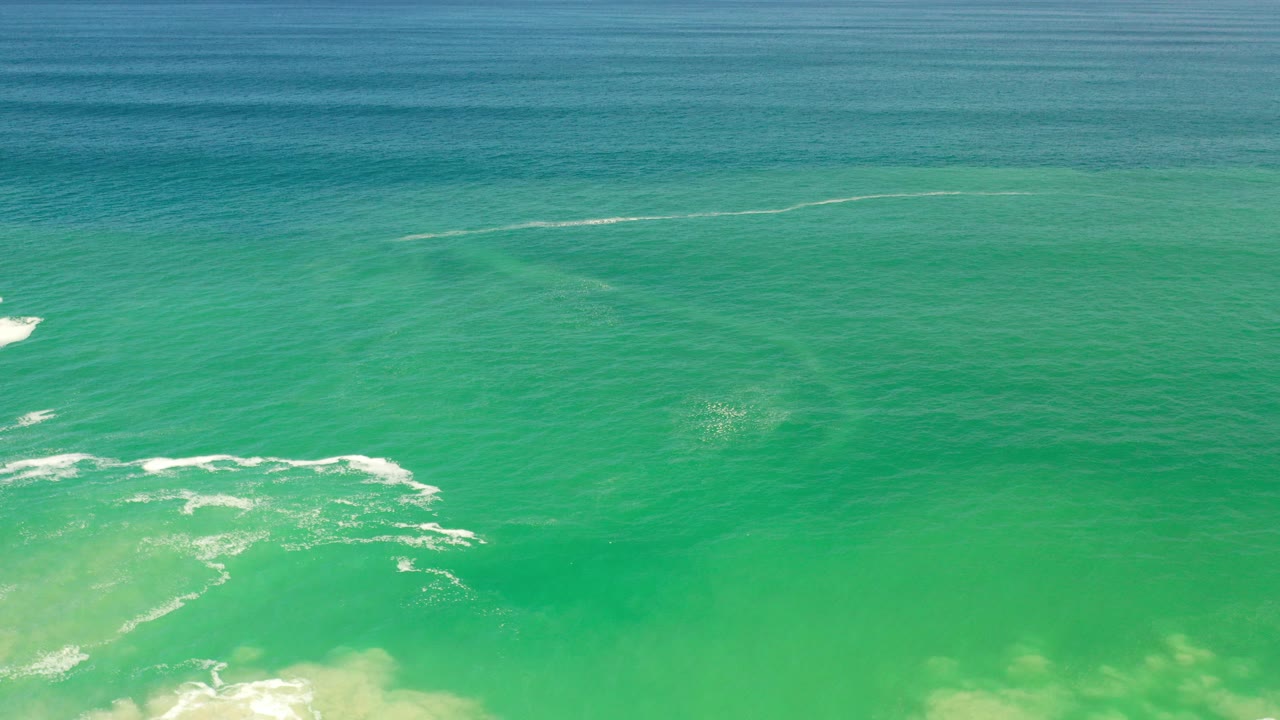 Blue-green ocean water in Los Cabos, Mexico with scattered white foam near the lower left, darker underwater patches, a blue horizon band, and no visible people.