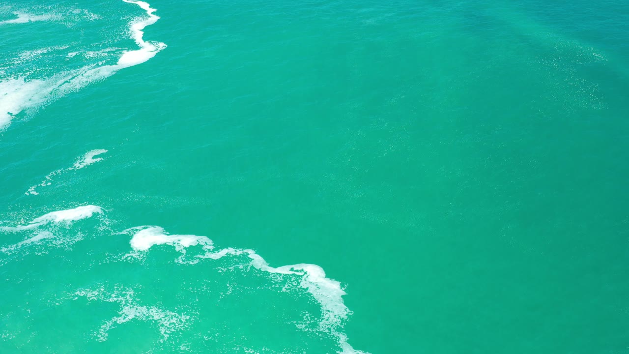 Overhead view of turquoise-green ocean water in Los Cabos, Mexico, with white surf breaking along the upper left and lower right edges and no visible people.