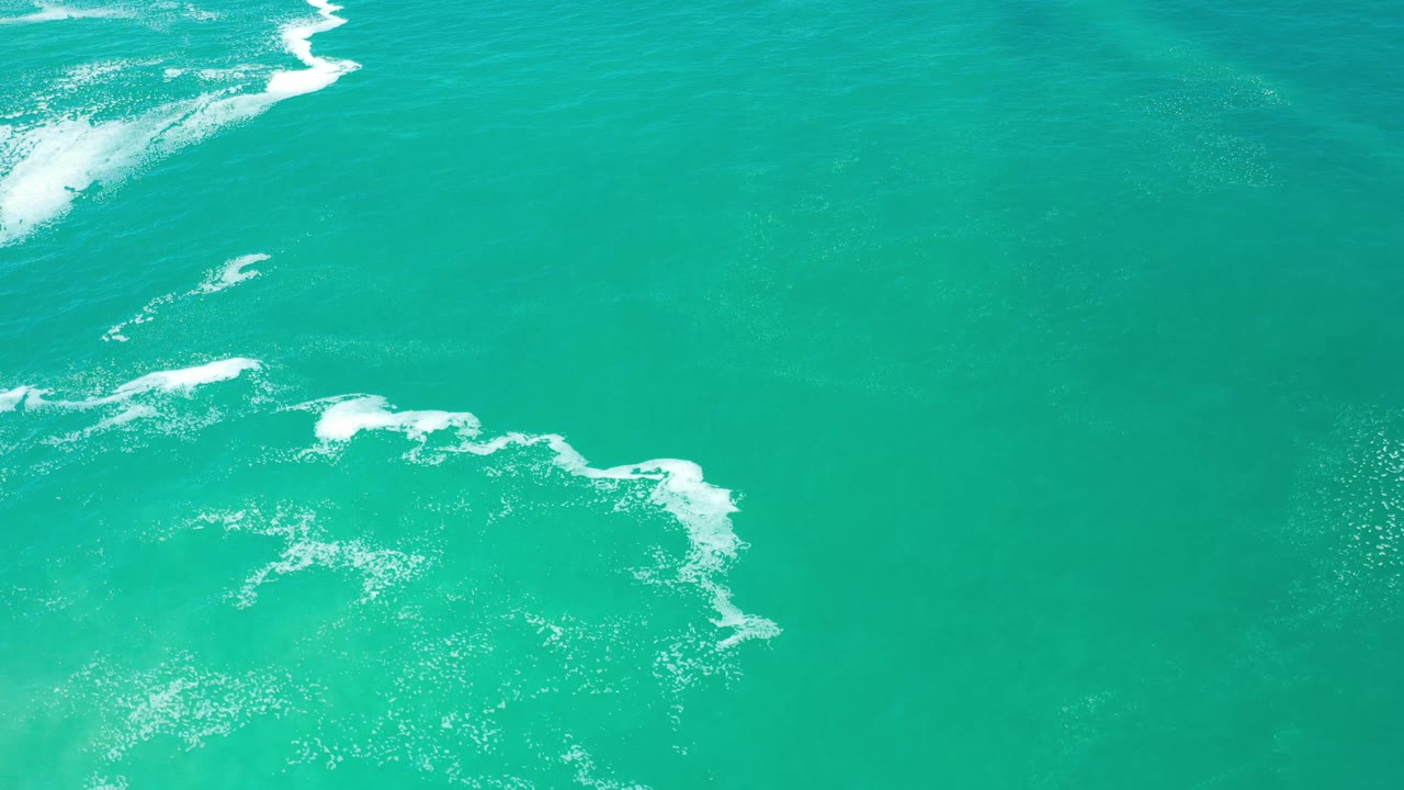 Aerial frame of blue-green water in Los Cabos, Mexico, with a brighter foam patch near the upper left and broken white foam through the middle, no visible people.