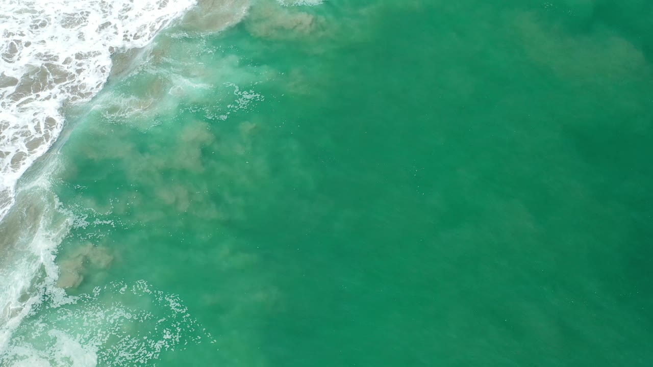 Overhead Los Cabos frame showing green seawater, scattered foam bubbles, and cloudy tan sand patches beneath the surface, with no visible people.