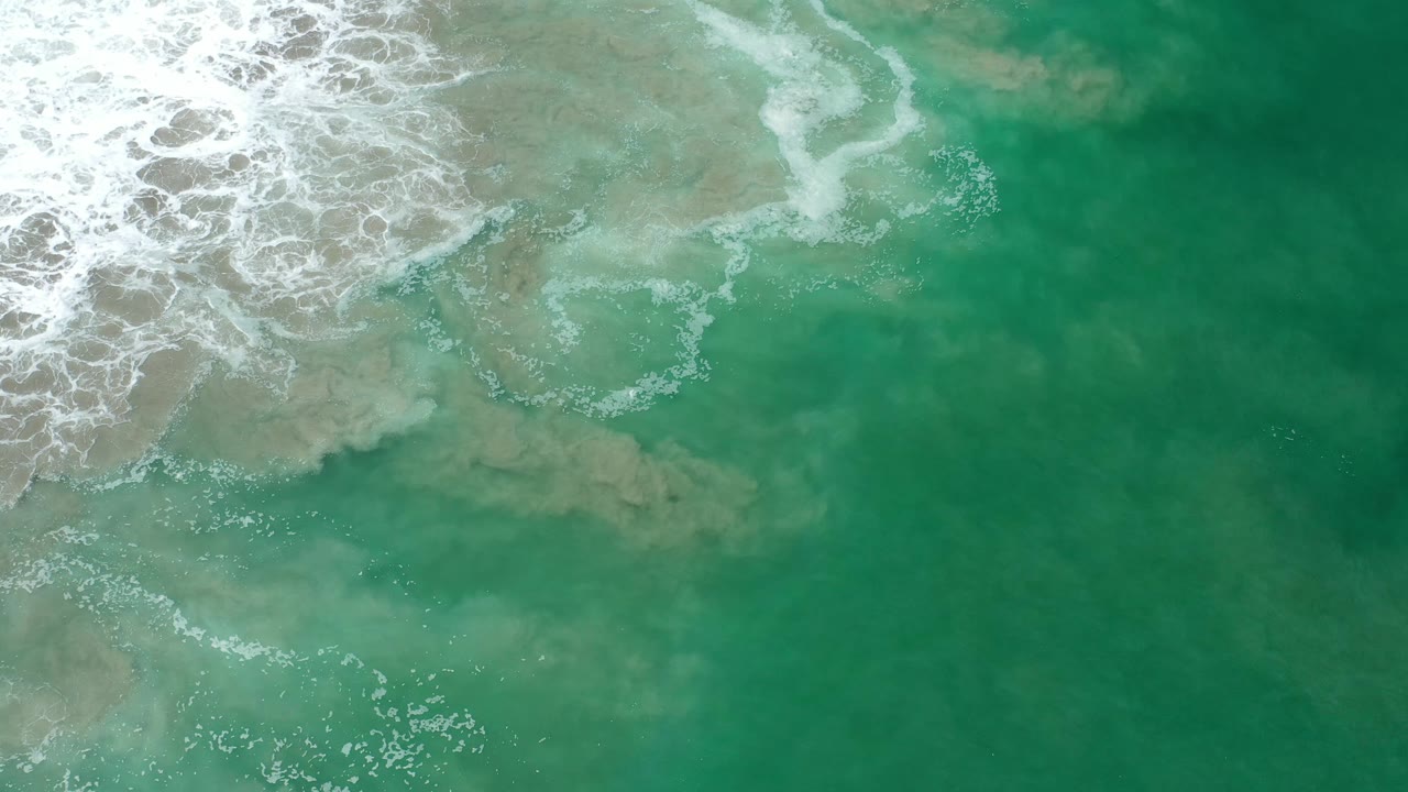 Overhead Los Cabos sea view with white surf curling along the upper left edge and green water filling the lower frame, no visible people.
