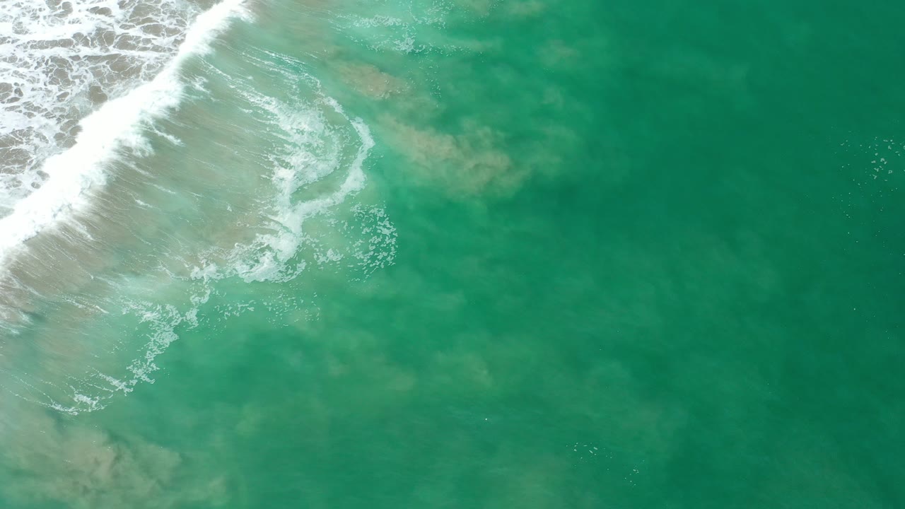 Overhead view of blue-green water in Los Cabos, Mexico, with white surf foam entering from the upper left and pale underwater patches visible, no visible people.