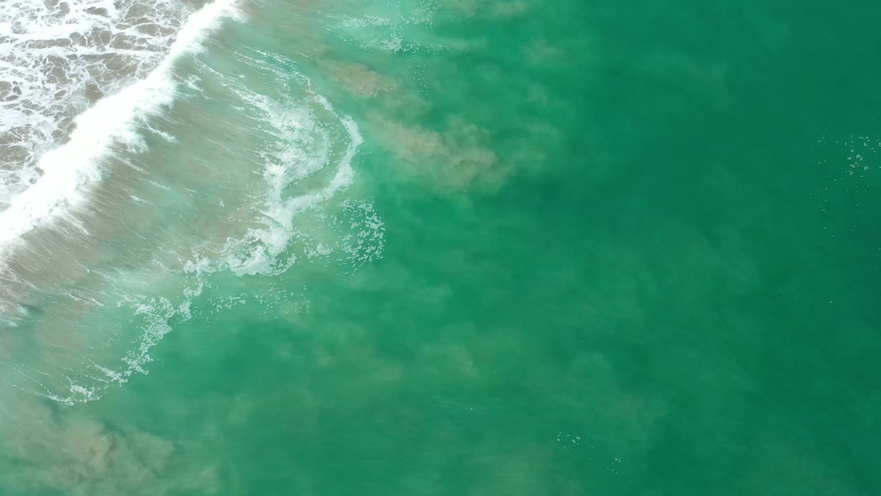 Aerial frame over Los Cabos water where a curved strip of white wave foam cuts diagonally across translucent green water, no visible people.
