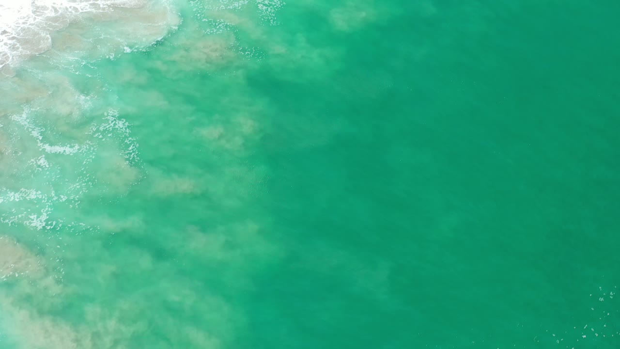 Overhead view of turquoise water in Los Cabos, Mexico, with pale underwater patches and white surf entering from the upper left, no visible people.