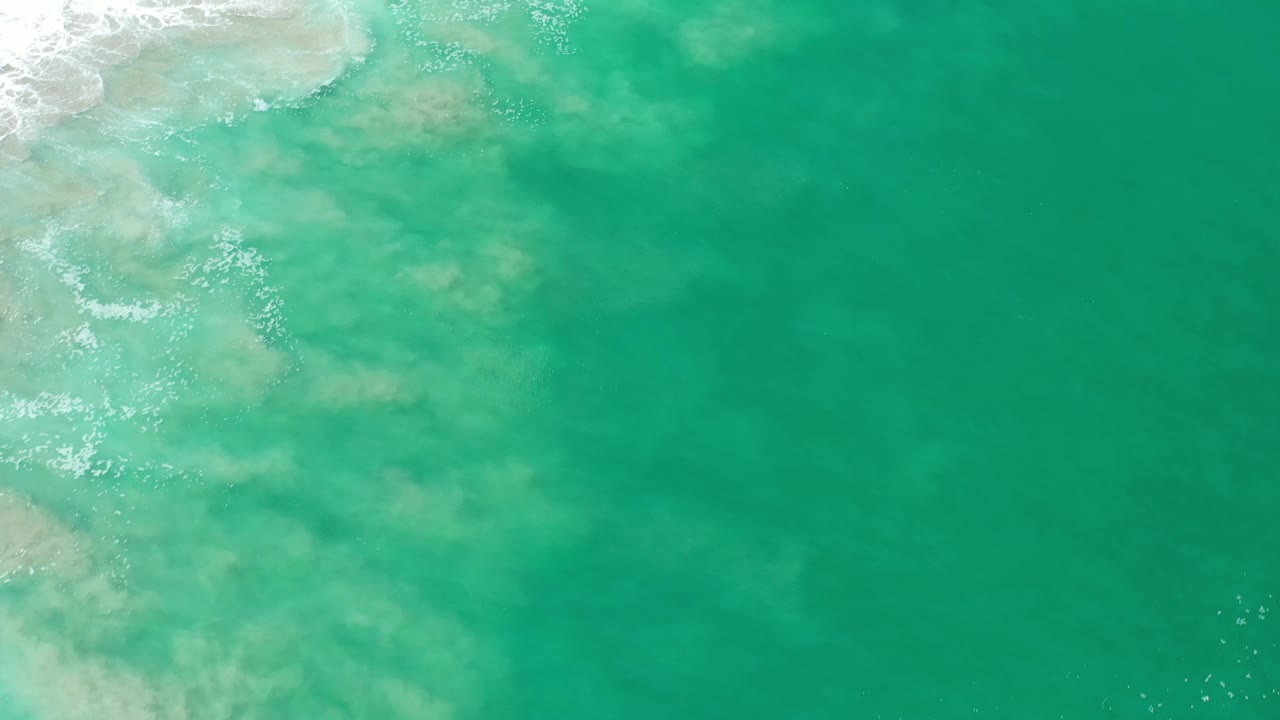 Top-down Los Cabos ocean frame with green turquoise water, faint sandy or rocky areas below the surface, and a small band of foam near the upper left, no visible people.