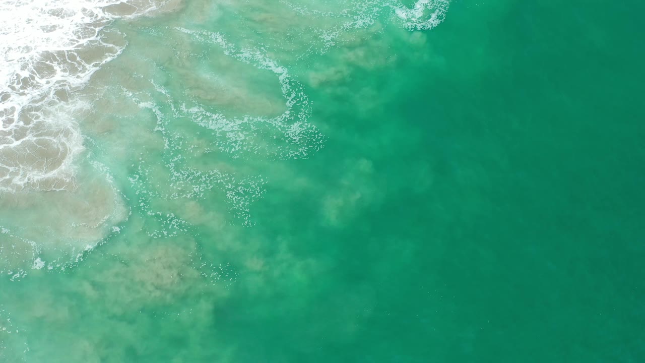 Green water fills most of the overhead frame in Los Cabos, Mexico, while broken white foam drifts away from the sandy shallows at left, with no visible people.