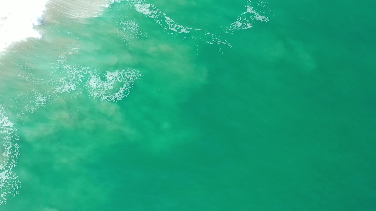 Overhead view of green-blue ocean water over pale sand in Los Cabos, with white surf entering at the upper left and no visible people.