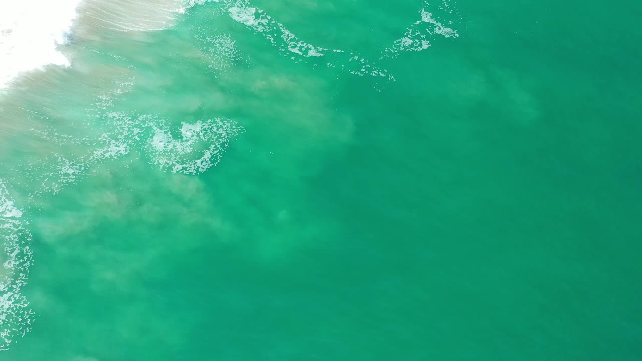 Overhead Los Cabos beach water frame with a foamy white break along the upper left, translucent green water, pale sand below, and no visible people.