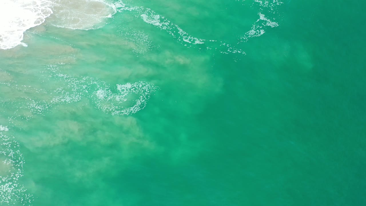Overhead view of teal ocean water in Los Cabos, Mexico, with pale surf wash and foam entering from the upper left, no visible people.