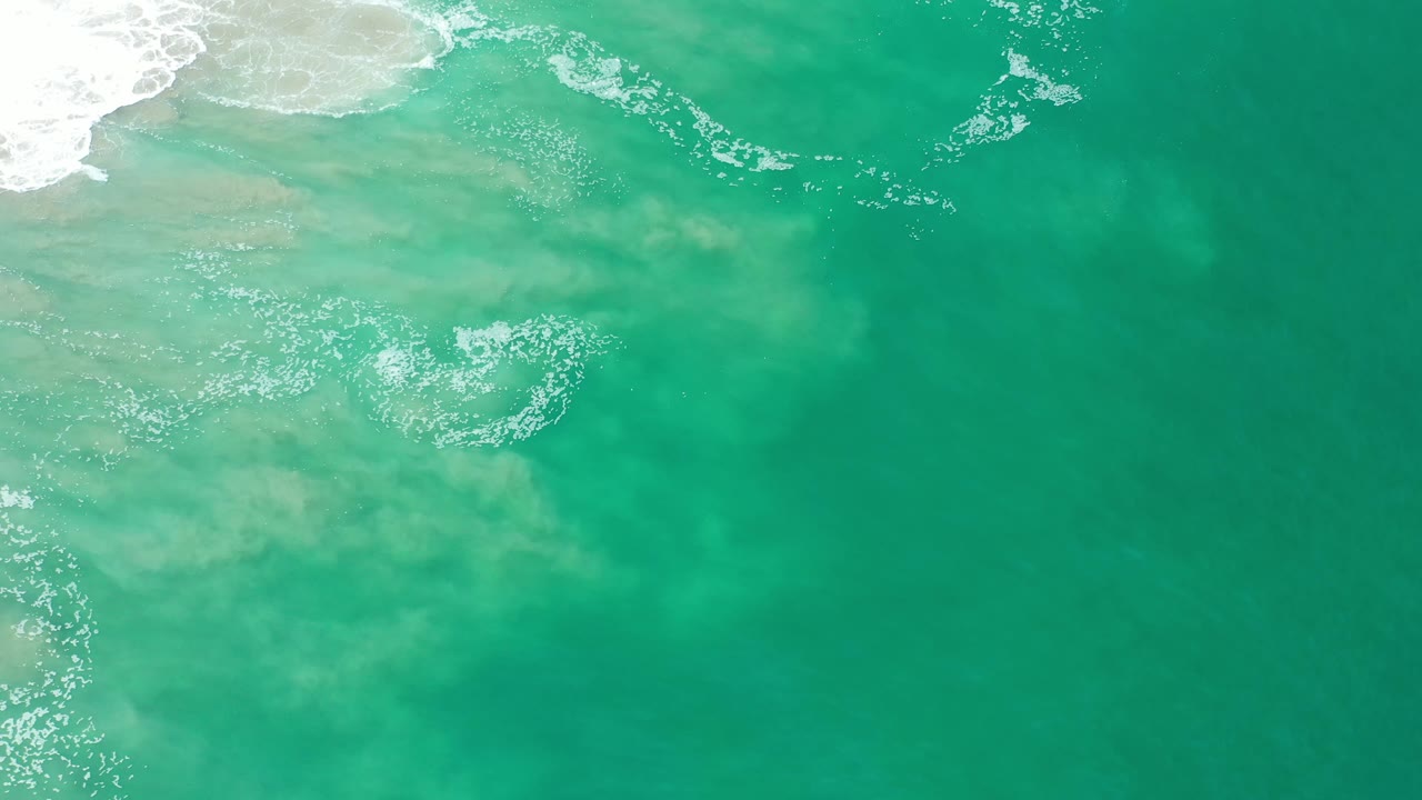 Overhead Los Cabos ocean frame with teal water, scattered foam flecks, and a pale sand-colored area just below the upper-left surf edge, no visible people.