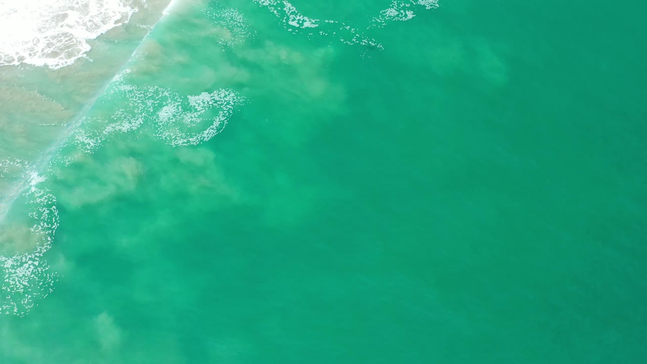 Aerial Los Cabos shoreline frame with white surf pushed farther down the left edge, pale sand visible under the water, and no visible people.