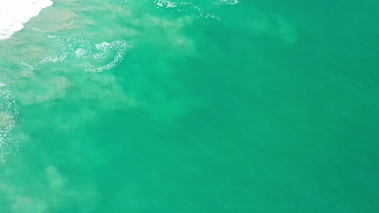 Overhead view of clear green-blue water in Los Cabos, Mexico, with pale sand under the surface, white surf entering from the upper left, and no visible people.