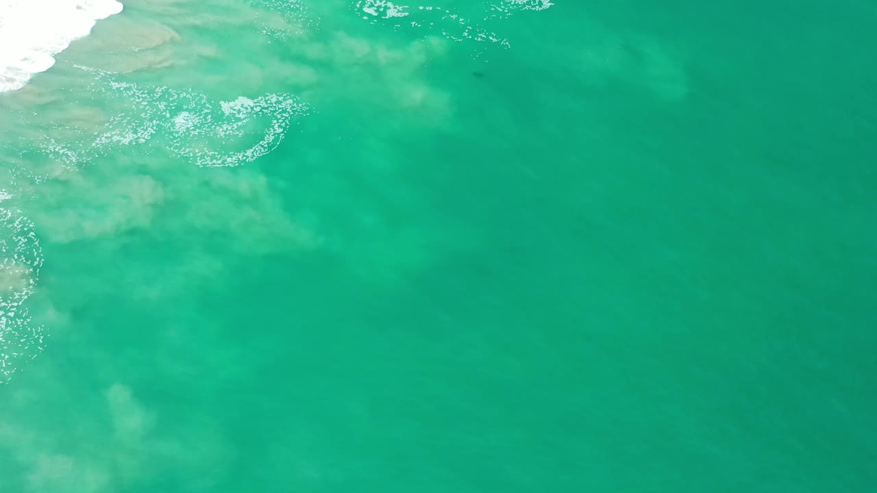 Aerial-looking frame of aquamarine water in Los Cabos, Mexico, with scattered white foam near the top edge, sandy shallows below, and no visible people.