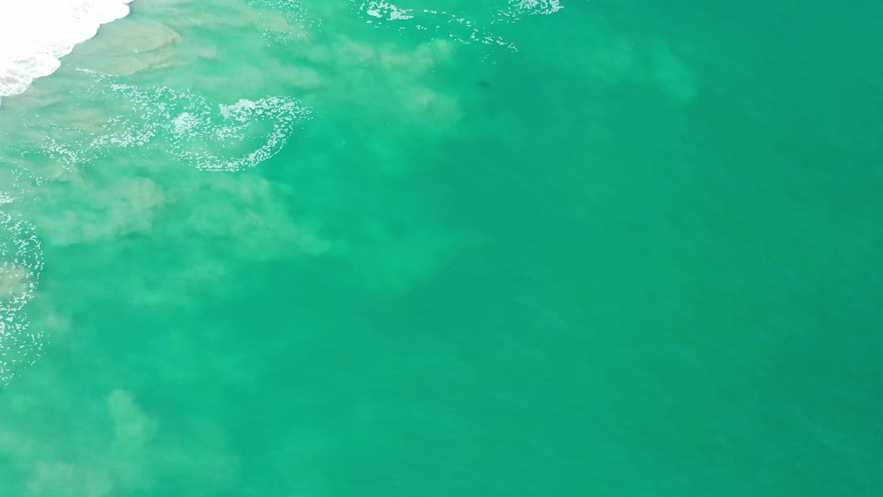 Clear turquoise ocean surface in Los Cabos, Mexico, seen from above with foamy shore wash at the upper left and open water across the right side, no visible people.