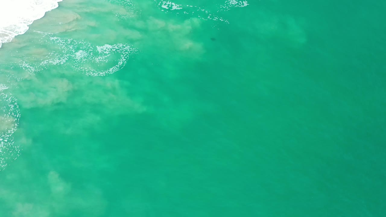 Green ocean water in Los Cabos, Mexico, with pale sand patches visible under the surface, surf foam along the left edge, and no visible people.