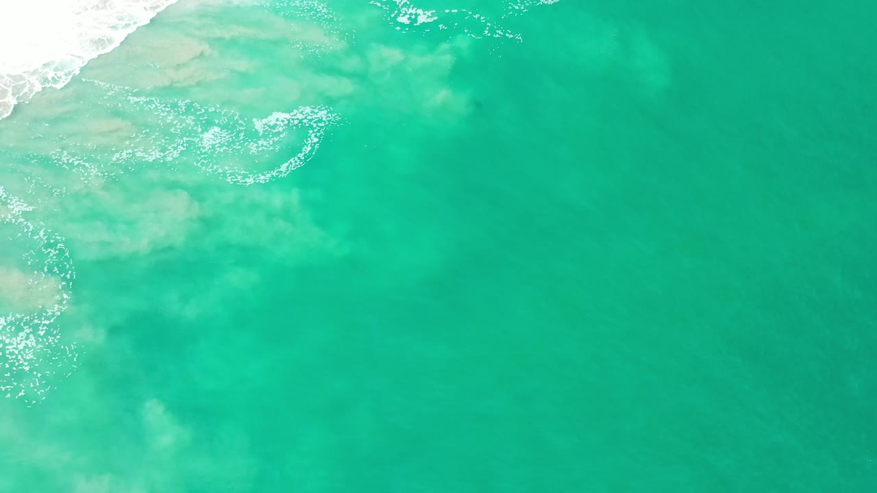 Overhead view of turquoise-green water in Los Cabos, Mexico, with white surf entering from the upper-left edge and no visible people.