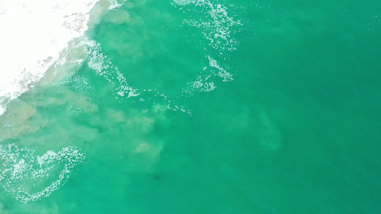 Overhead view of blue-green ocean surf in Los Cabos, Mexico, with white foam along the upper left edge and no visible people.