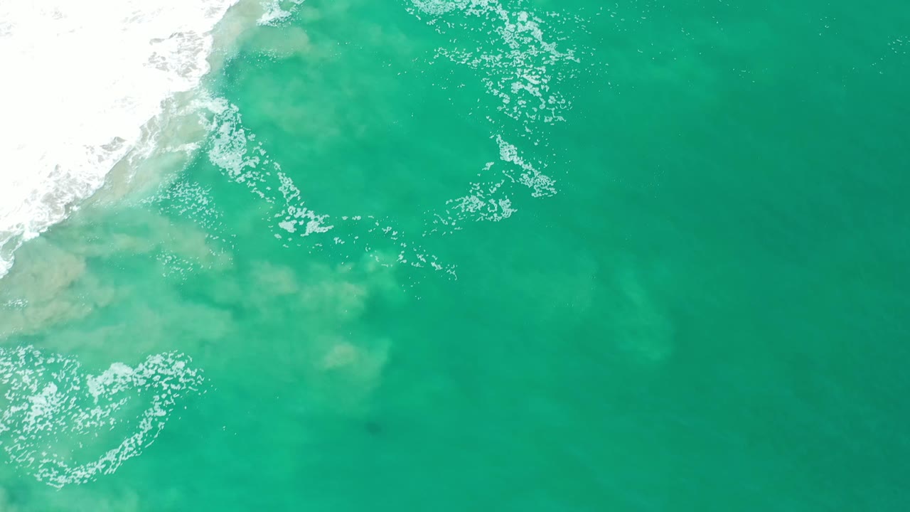 Aerial frame over Los Cabos surf where white foam gathers near the top left while pale sand clouds spread under the turquoise water, with no visible people.