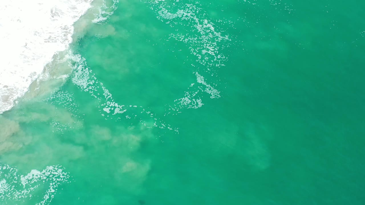 Overhead blue-green ocean with white foam and cloudy sand patches.