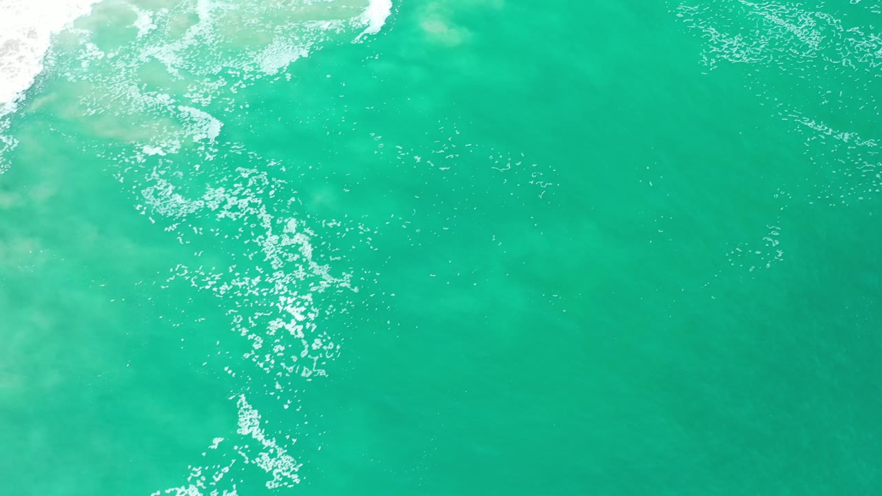Overhead view of green-turquoise ocean in Los Cabos, Mexico, with white surf foam entering near the top left and no visible people.