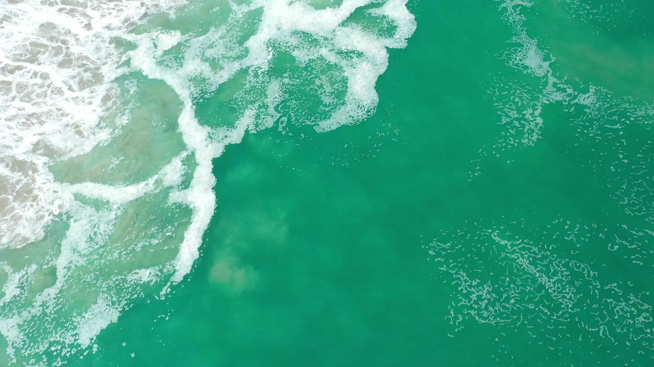 Overhead view of blue-green ocean water in Los Cabos, Mexico, with white foam breaking from the upper left and no visible people.