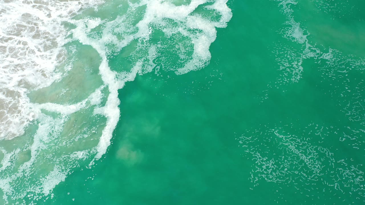 Overhead Los Cabos ocean frame with a larger band of white foam curling across the top and clear green water below, with no visible people.